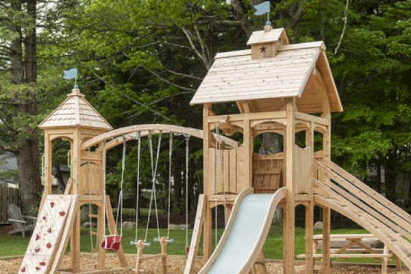 The image shows a wooden playground set with a slide, swings, a rock climbing wall, and two towers connected by a bridge, surrounded by trees.