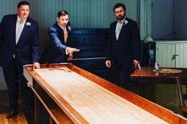 Three men in suits are playing shuffleboard in a room with hardwood floors and wood paneling. One man is in the act of sliding a puck.