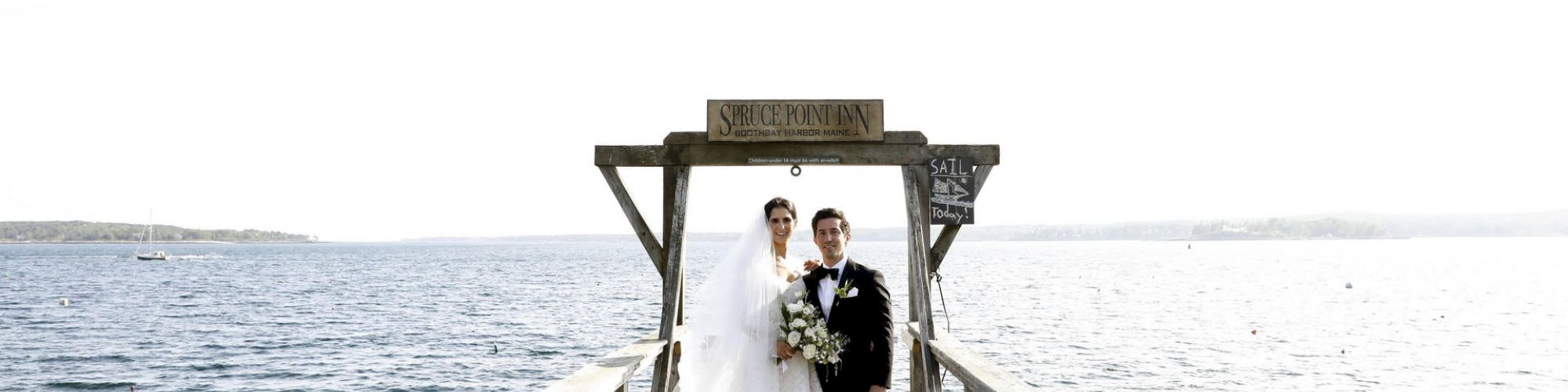 A bride and groom standing on a dock by a large body of water, with a few kayaks visible in the background, under a wooden structure.