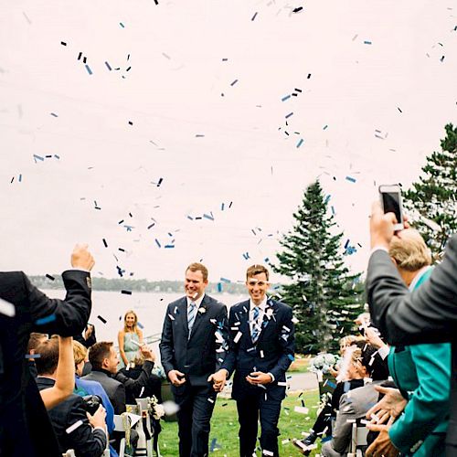 Two people in formal attire are walking down an outdoor aisle, surrounded by confetti and guests, with a lake and trees in the background.