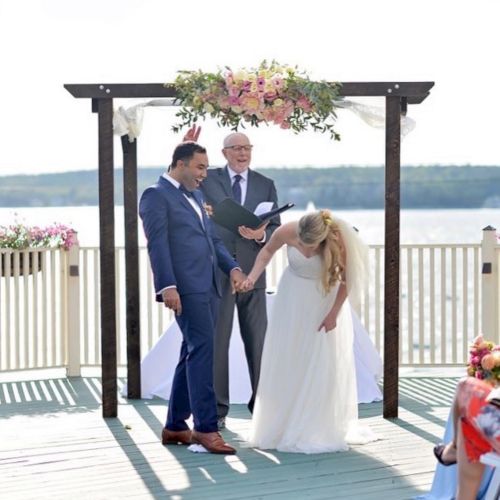 A couple is holding hands under a floral arch at a waterfront wedding ceremony, while an officiant stands behind them, smiling.