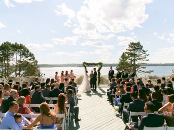 A wedding ceremony set outdoors near a body of water with guests seated, the couple standing under an arch, and the bridal party beside them.