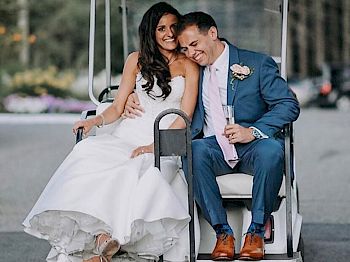 A bride and groom are sitting on a golf cart, both smiling and appearing joyful. The bride is in a white dress, and the groom is in a blue suit.