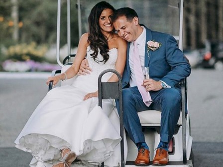 A bride and groom are sitting on a golf cart, both smiling and appearing joyful. The bride is in a white dress, and the groom is in a blue suit.