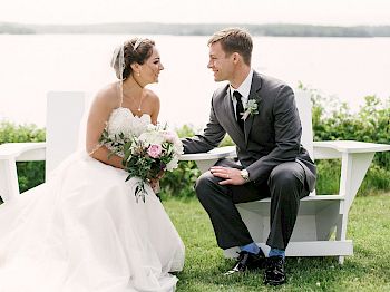 A bride and groom share a joyful moment, seated on a white bench near a scenic lake, with greenery in the background, both smiling happily.