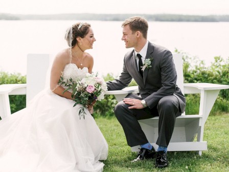 A bride and groom share a joyful moment, seated on a white bench near a scenic lake, with greenery in the background, both smiling happily.