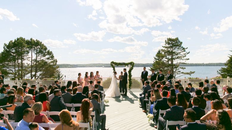 A wedding ceremony is taking place outdoors by a lake, with guests seated and the couple standing under an arch.