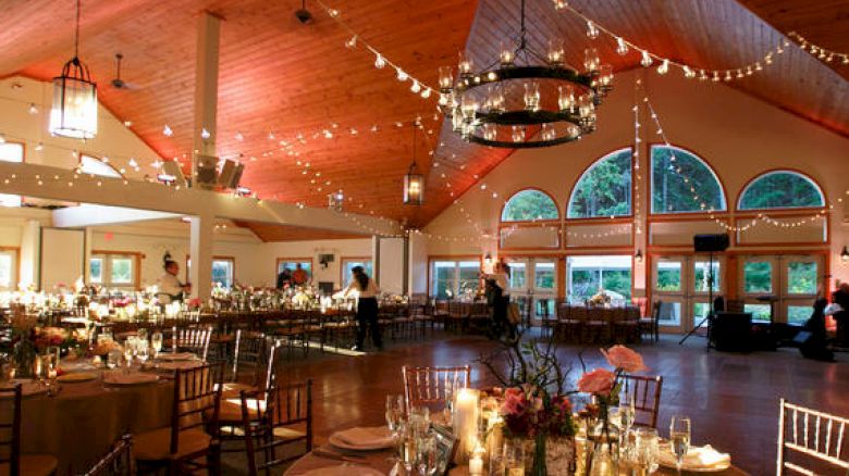 A beautifully decorated event space with round tables set for dining, string lights hanging from the wooden ceiling, and large windows in the background.