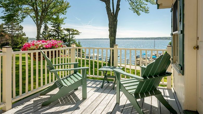Two green Adirondack chairs on a wooden deck overlook a scenic view of a lake with trees and flowers in the background, under a clear sky.