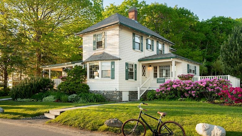 A charming two-story house with a bicycle in front, surrounded by vibrant greenery and colorful flowers, featuring a wrap-around porch.