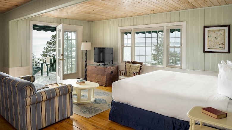This image shows a cozy bedroom with a large bed, TV, sofa, and a view of the ocean from a balcony with outdoor seating.