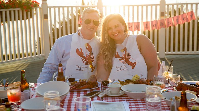 A couple wearing lobster bibs smiles at an outdoor table set for a meal, with drinks and food on a checkered tablecloth.