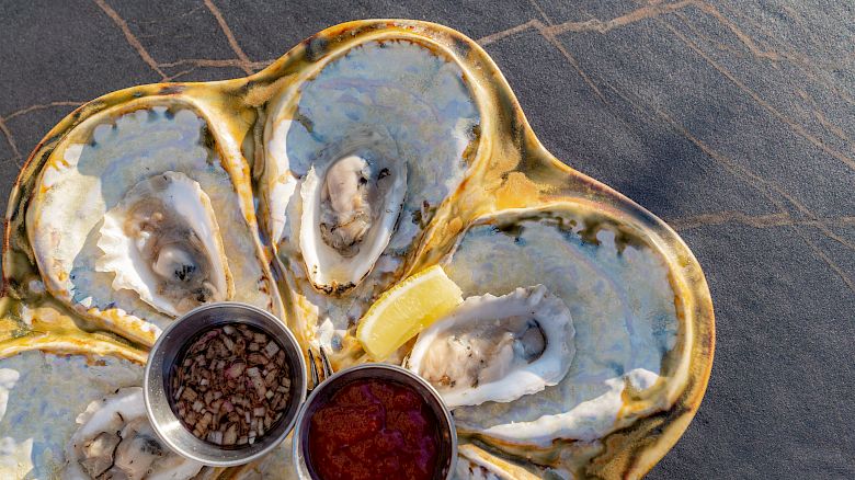A plate of oysters with two small condiment cups containing toppings and lemon wedges, placed on a dark surface.