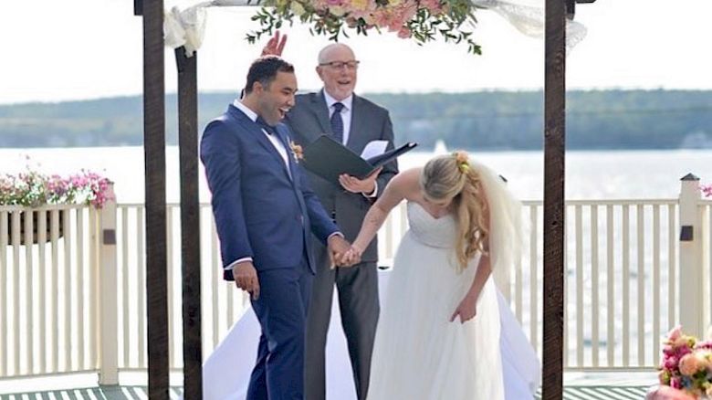A newlywed couple holds hands and smiles under a floral arch by a lakeside, with an officiant standing behind them, during an outdoor wedding ceremony.