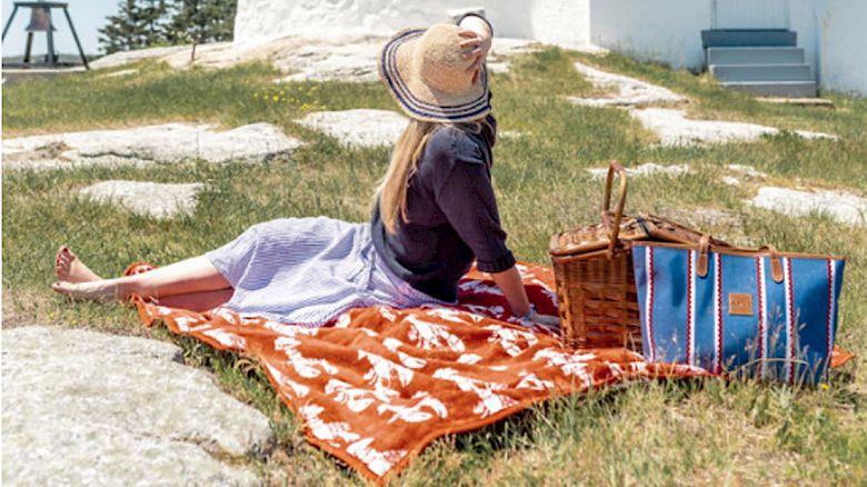 A person sits on a blanket having a picnic near a lighthouse on a bright, sunny day, surrounded by green grass and clear skies.