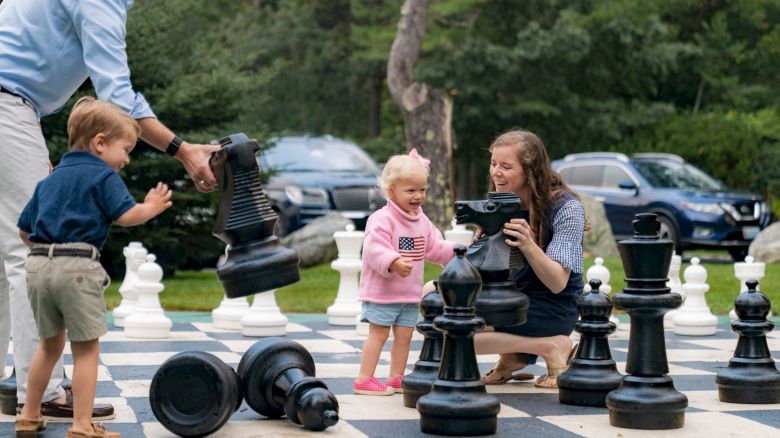 A family is playing with oversized chess pieces on an outdoor board. The young children and adults seem to be enjoying the activity.