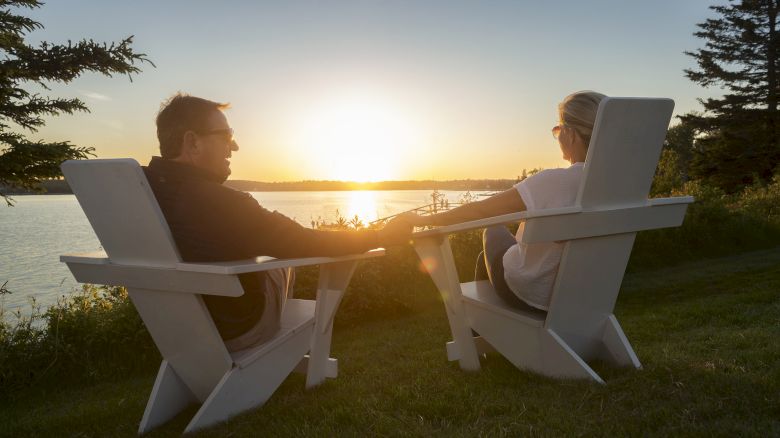 Two people sit in Adirondack chairs holding hands and watching a sunset over a serene body of water, surrounded by trees and greenery.