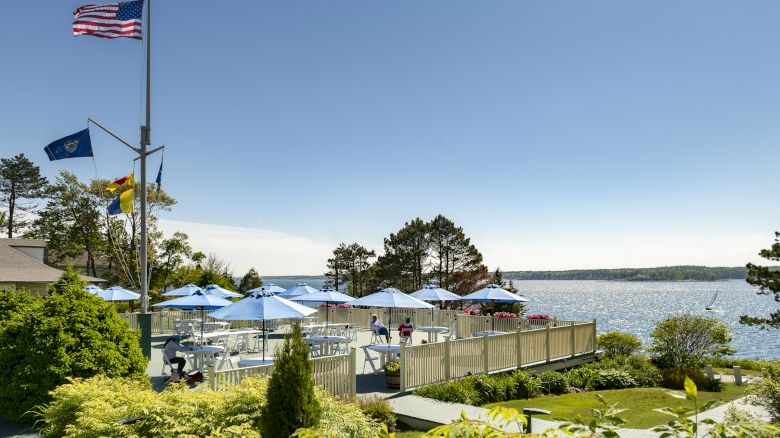 An outdoor seating area by a lake with blue umbrellas, surrounded by lush greenery and flags, all under a clear blue sky.