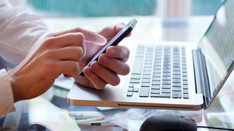 A person is using a smartphone while sitting in front of an open laptop computer on a desk.