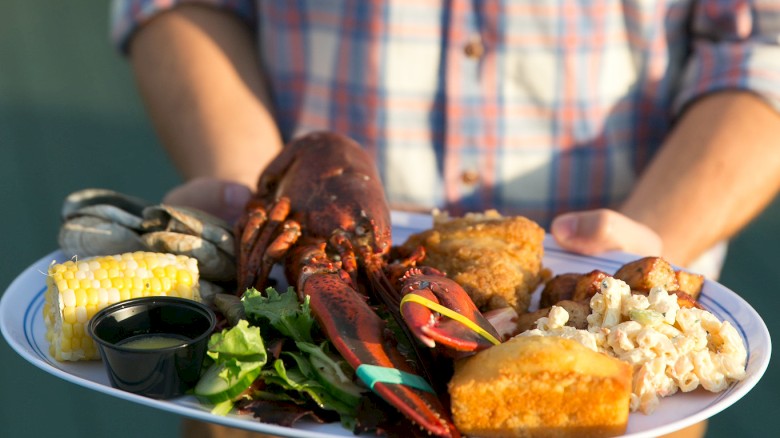 A person in a plaid shirt is holding a plate with a lobster, corn, greens, bread, and other assorted foods.