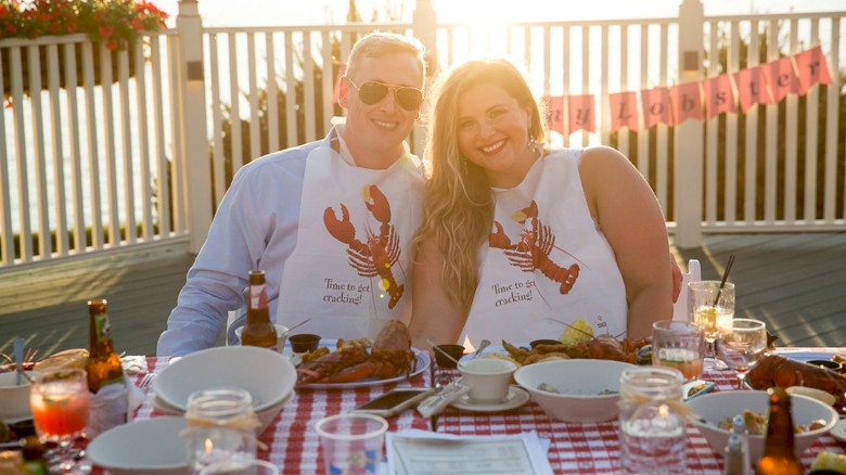 A smiling couple wearing lobster bibs sit at a table with a checkered tablecloth, enjoying a seafood meal outdoors.