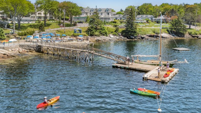 The image shows people kayaking near a wooden dock with a small sailboat, located by a waterfront community with houses and greenery in the background.