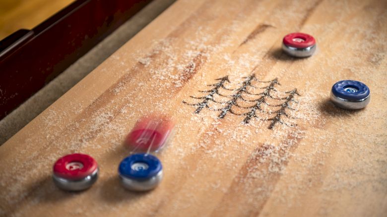 The image shows a game of table shuffleboard with red and blue pucks sliding on a board dusted with powder. Ending the sentence.