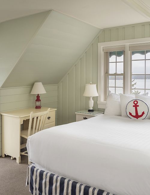 A cozy bedroom with white and light green decor, nautical-themed accents, a bed with an anchor pillow, and a view of the water through windows.
