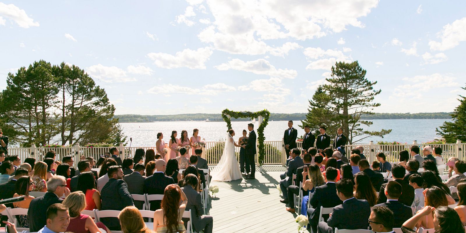 A wedding ceremony is taking place outdoors with guests seated, officiant, wedding party, and couple under an arch by a scenic lake.