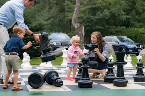 A family plays with giant chess pieces on an oversized chessboard outdoors, with children excitedly interacting with the large pieces.