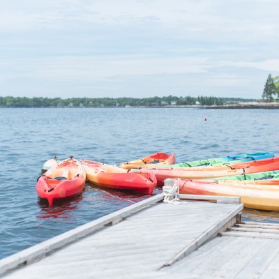 Several colorful kayaks are tied to a wooden dock by calm water with a distant shoreline and trees under a cloudy sky.