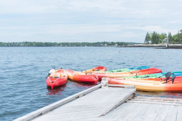 Colorful kayaks are lined up and tied to a dock on a calm lake with trees and houses visible in the background.
