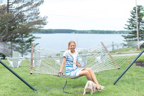 A woman in a striped dress sits on a hammock with a small dog in front of her, near a lake with trees in the background, ending the sentence.
