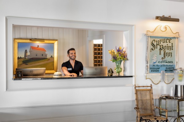 A man is standing behind a reception desk with a painting and flowers. An antique sign, 