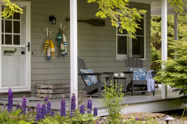 A cozy porch with two Adirondack chairs, colorful life jackets hanging on the wall, surrounded by lush greenery and blooming flowers, creating a serene setting.