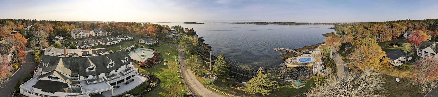 A panoramic view of a coastal resort featuring buildings, a swimming pool, trees with fall foliage, a body of water, and roads.