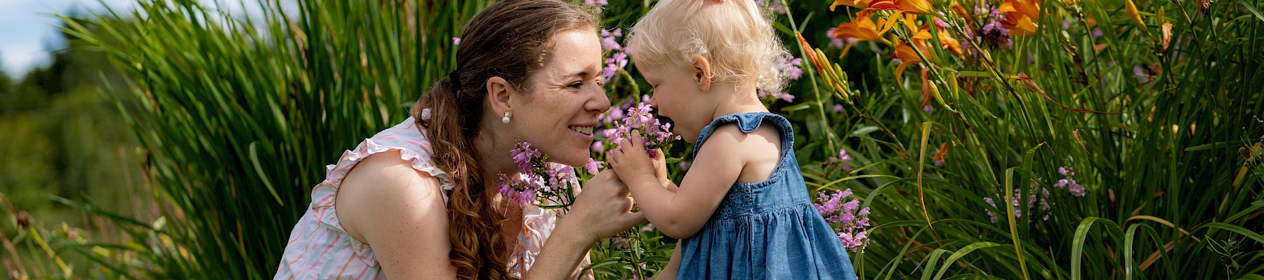 A woman and a child are smelling flowers in a garden, surrounded by lush greenery and blooming orange lilies.