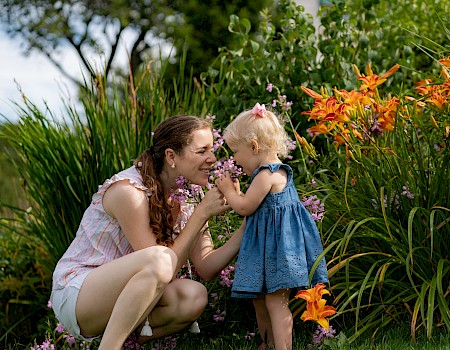 A woman and a child are smelling flowers in a garden, surrounded by lush greenery and blooming orange lilies.