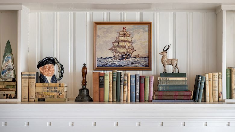 A bookshelf with assorted books, a ship painting, a sailor bust, a deer figurine, and other decorative items displayed on a white wooden mantel.