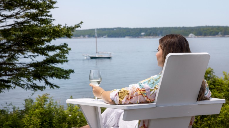 A person sits in a white Adirondack chair, holding a wine glass, and looks out at a scenic lake with a boat in the distance and trees on the shore.