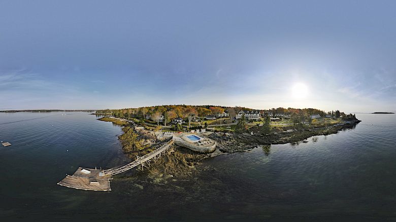 A panoramic view of a coastal area with a dock, buildings, and trees under a clear sky with the sun shining brightly in the background.