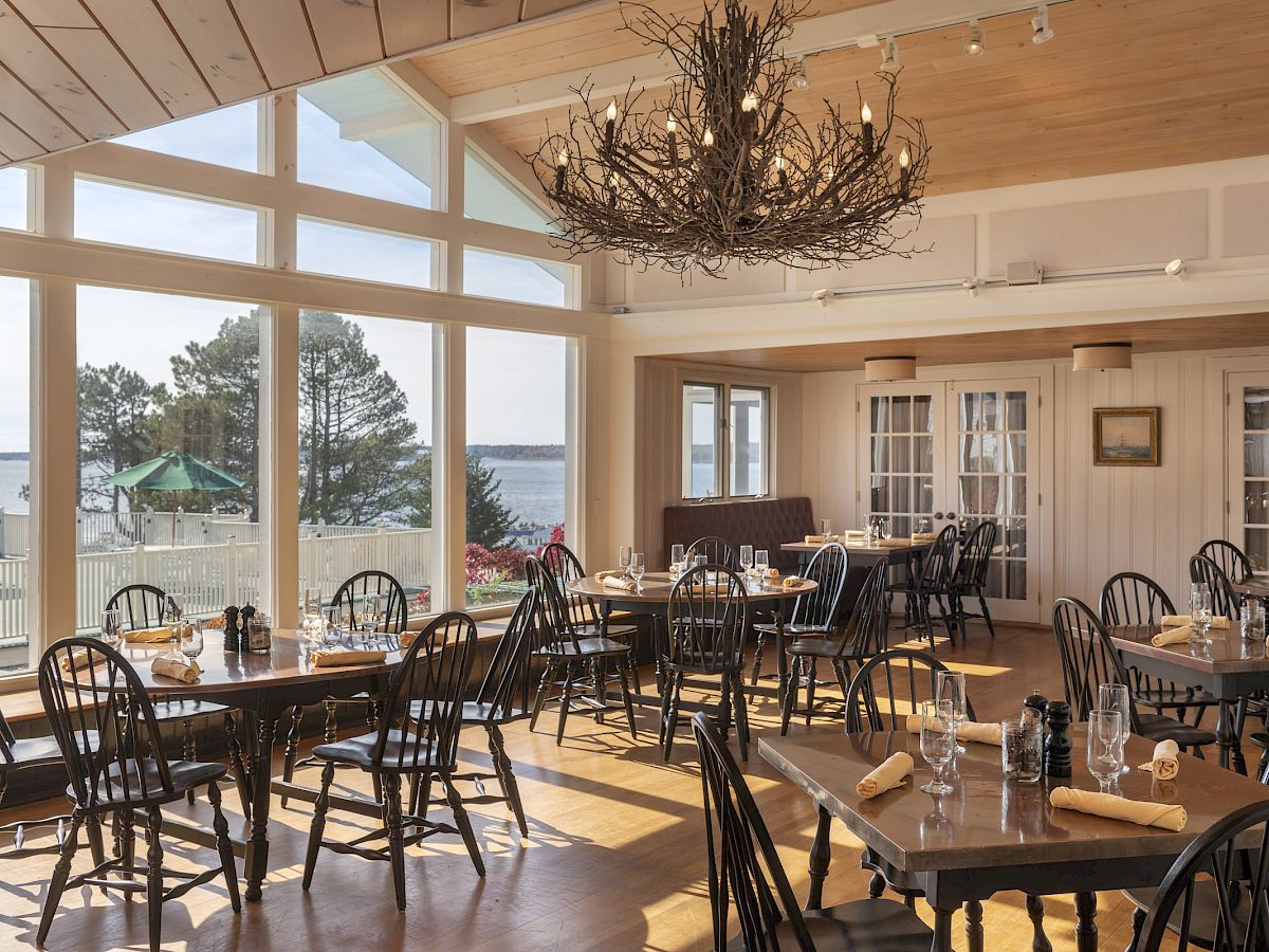 A cozy restaurant with wooden tables and chairs, large windows offering a scenic view of water and trees, and a rustic chandelier on the ceiling.
