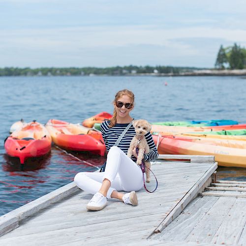 A woman sits on a dock holding a small dog; colorful kayaks are in the background, and there is a lake with trees on the horizon.