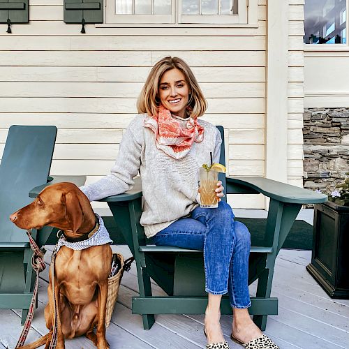 A woman sits on a porch chair with a drink in hand, smiling, while a dog sits beside her, both in a relaxed, cozy setting.