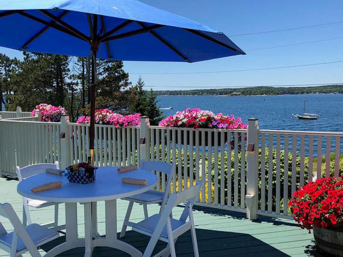 A patio with a white table and chairs, blue umbrella, overlooking a lake with boats and surrounded by colorful flowers and plants.