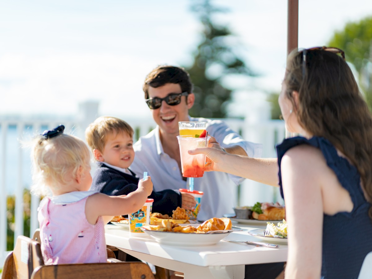 A family enjoys a meal at an outdoor table, with two adults and two children sharing food and drinks, smiling and toasting each other.