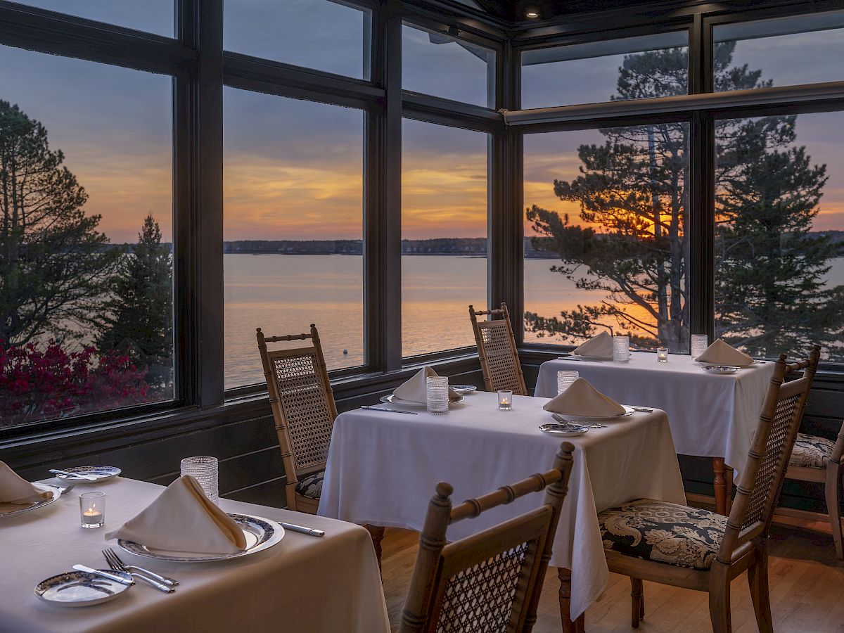 A cozy restaurant with tables set for dining, large windows offering a stunning sunset view over a body of water, and a few trees visible outside.