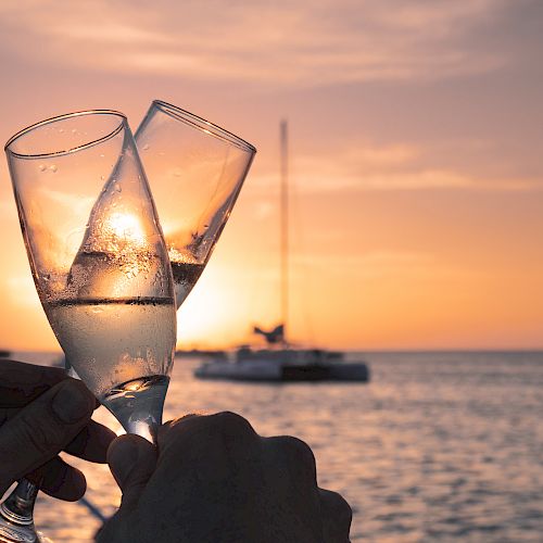 Two champagne glasses clink together against the backdrop of a serene sunset over a body of water with sailboats in the distance.