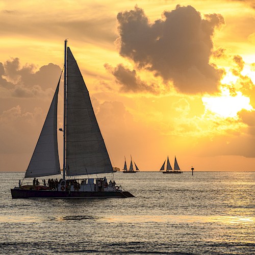 A sailboat on a calm ocean at sunset with other sailboats in the background and a dramatic sky filled with clouds, ending the sentence.