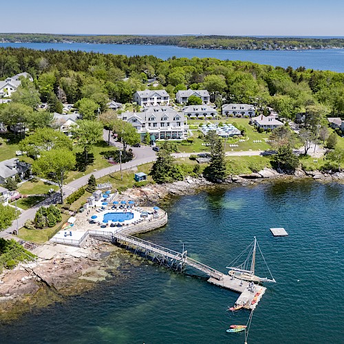 Aerial view of a waterfront resort with multiple buildings, a swimming pool, and a dock extending into a body of water surrounded by greenery.